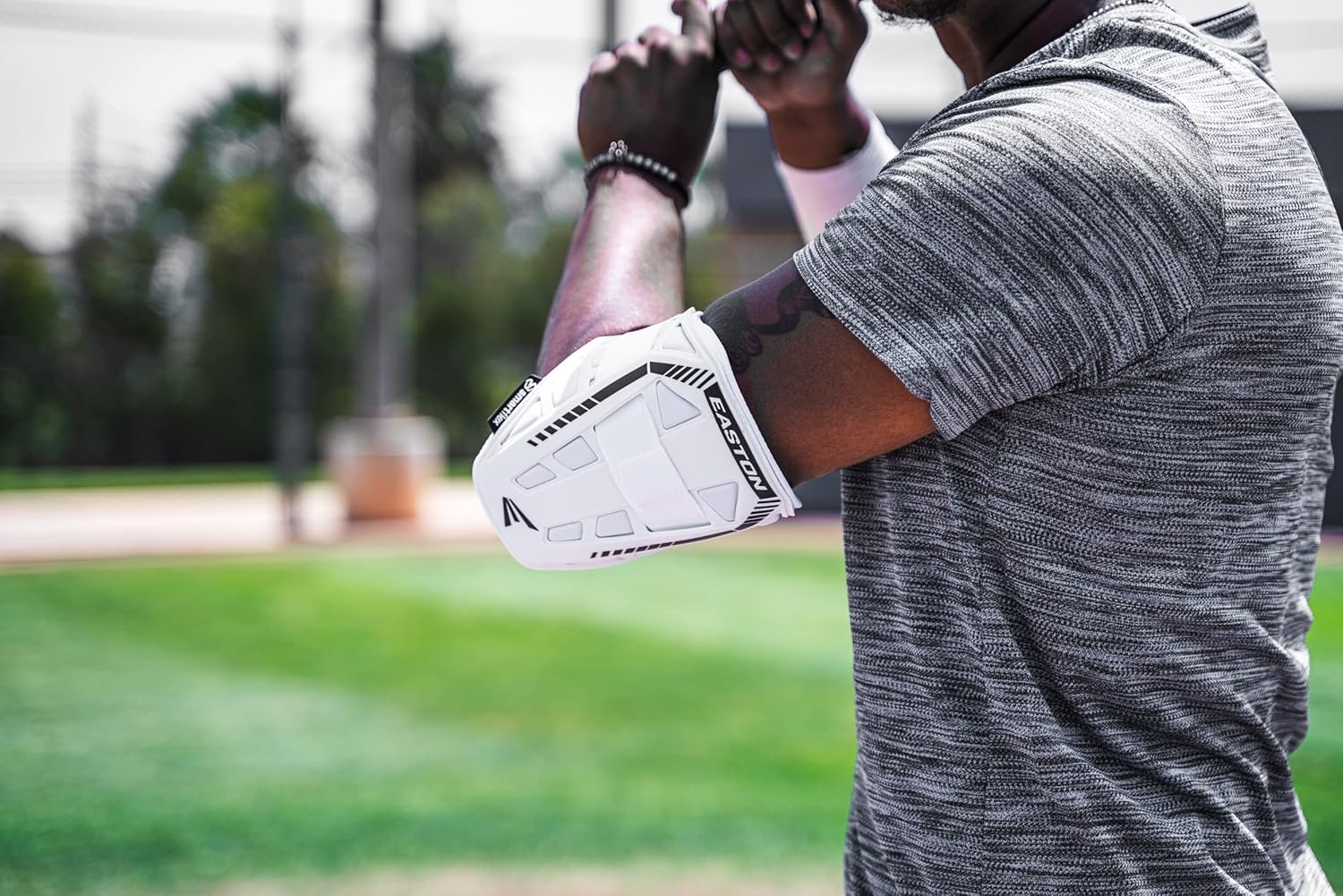 A person wearing a gray striped shirt and an Easton Compact Batter Elbow Guard: ES37 stands on a grassy field with arms raised, blurred trees in the background.