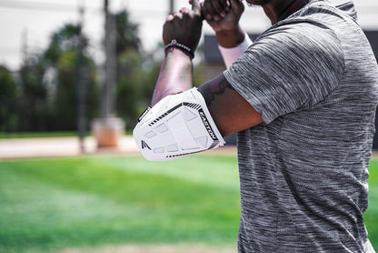 A person wearing a gray striped shirt and an Easton Compact Batter Elbow Guard: ES37 stands on a grassy field with arms raised, blurred trees in the background.