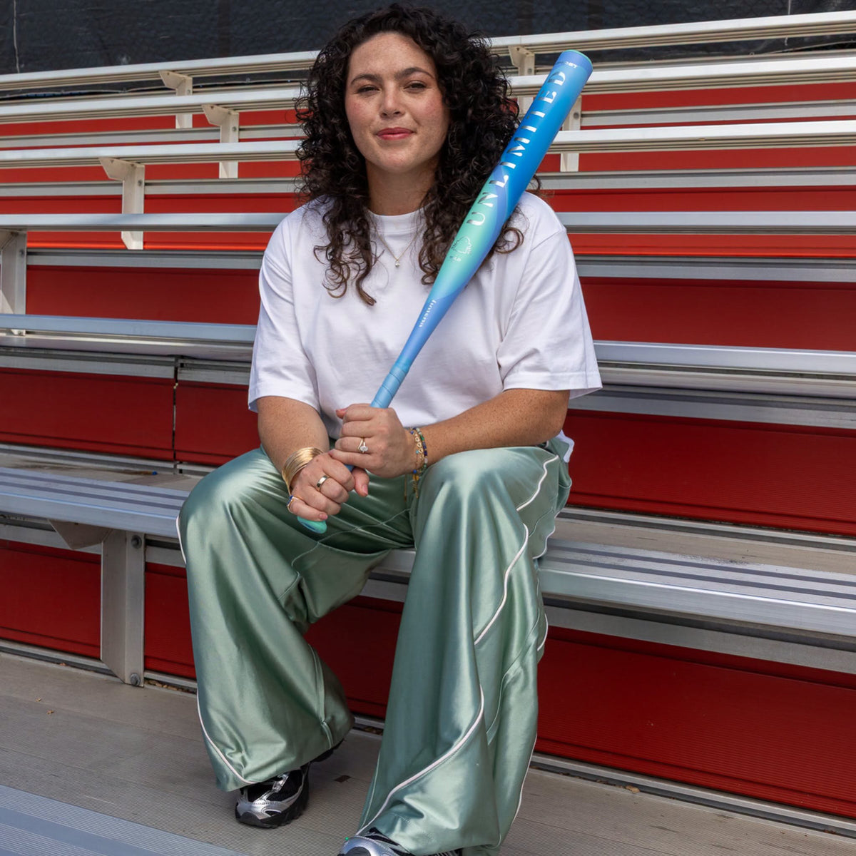 A woman sitting on red stadium bleachers holds a 2027 Easton Ghost Unlimited Wild (-10) Fastpitch Softball Bat: EFP6GUW10 across her lap.