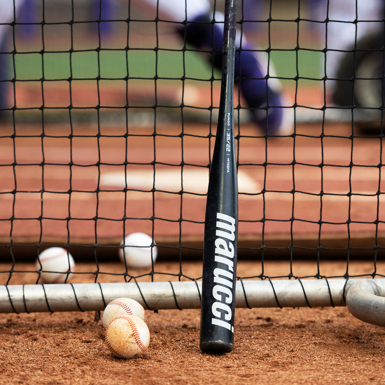 A Marucci Aluminum Fungo 35″ Training Bat: MTRBFA, crafted with high-strength alloy construction, rests against a net with its anti-vibration knob in view, as Marucci-branded baseballs are scattered on the field during practice.