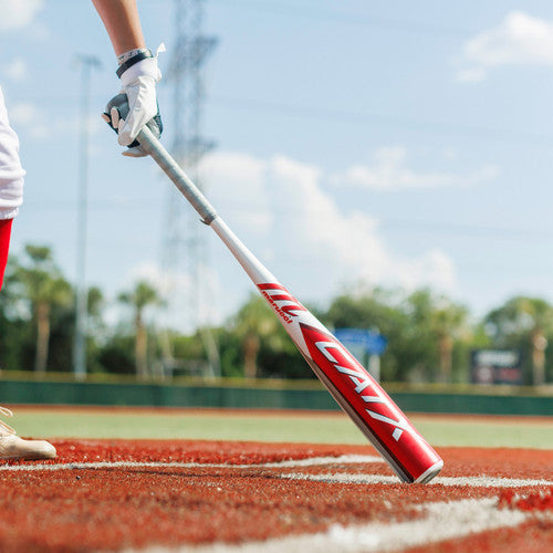 A baseball player in white and red gear holds a red and white CATX bat, standing on a baseball field with green grass, palm trees, and a blue sky in the background.