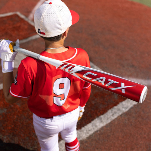 A young baseball player in a red jersey with the number 9 stands on a field, holding a CATX bat over his shoulder, facing away from the camera.