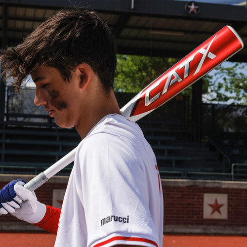 A young baseball player with black eye paint holds a red CATX bat over his shoulder, wearing a white jersey with marucci on the sleeve, standing on a baseball field with bleachers in the background.