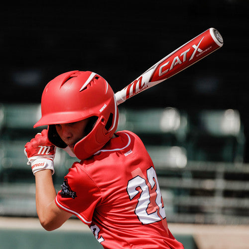 A young baseball player in a red uniform and helmet, holding a bat and preparing to swing, stands at home plate with the number 22 visible on their back. The background shows empty stadium seats.