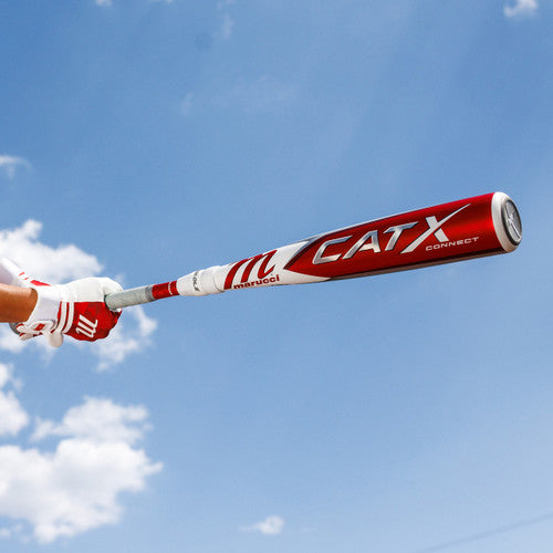 A person wearing red and white batting gloves holds a red and white Marucci CATX Connect baseball bat against a blue sky with a few clouds.