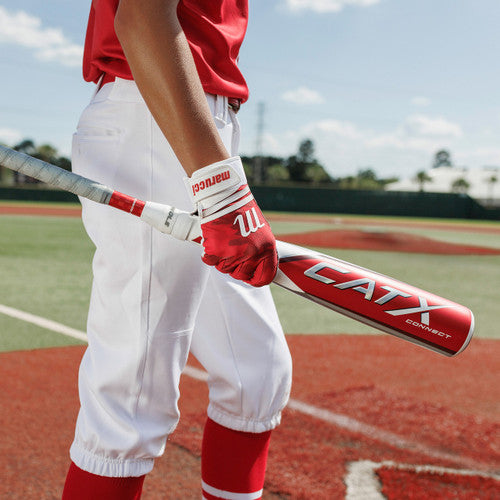 A baseball player in a red jersey and white pants stands on a field holding a red and white CATX Connect bat, wearing red gloves and striped socks. The field has green and red turf.