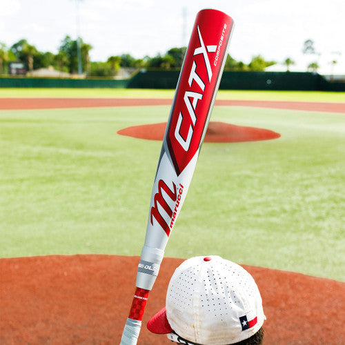 A person wearing a white baseball cap stands on a baseball field, holding a red and silver Marucci CATX baseball bat over their shoulder. The infield and outfield grass are visible in the background.