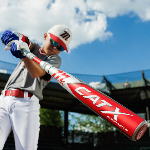 A young baseball player in a gray and red uniform swings a red and silver CATX Composite bat on an outdoor field under a partly cloudy sky.