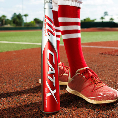 Close-up of a baseball player’s legs wearing red and white striped socks and red cleats, standing on a baseball field with a red Marucci CATX Composite bat resting on the ground.