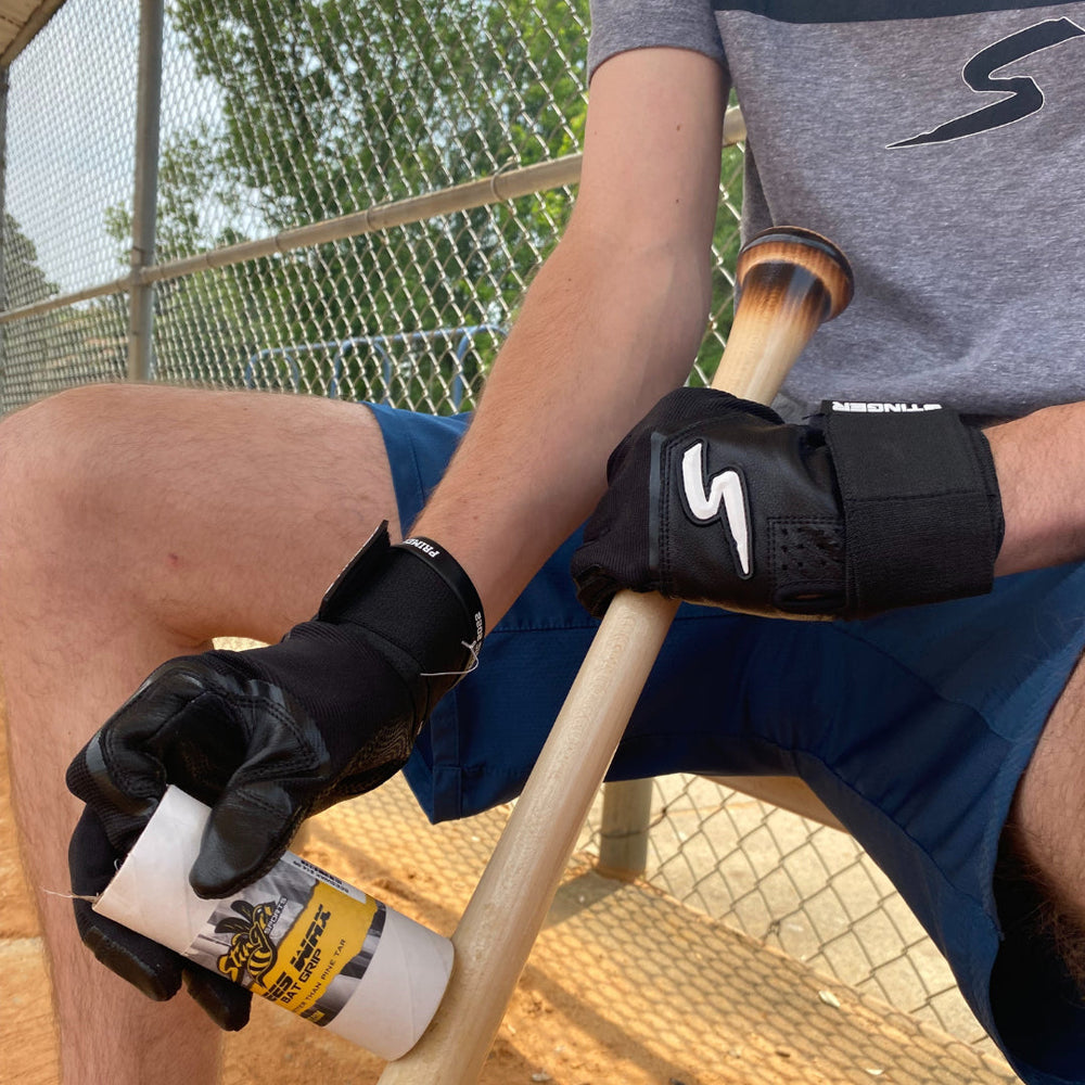 A person in black batting gloves and blue shorts sits on a bench in a baseball dugout, holding a bat and a Stinger Sports Stinger BEES WAX Bat Grip: BEESWAX. A chain-link fence and trees are visible in the background.