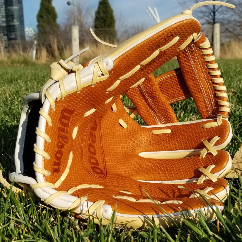 A tan and white Wilson baseball glove lies open on green grass outdoors, with trees and a building visible in the blurred background under a blue sky.