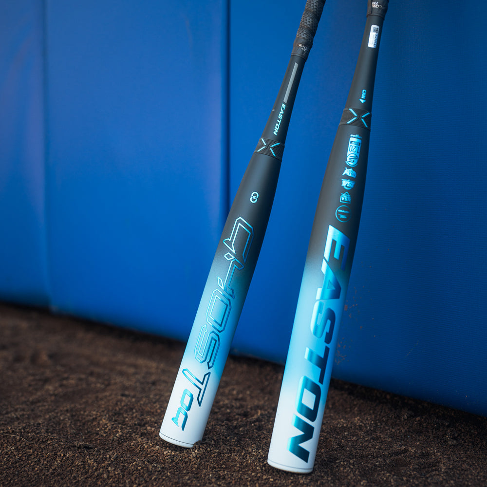 Two Easton Ghost OG (-11) 2025 Fastpitch Softball Bats by Easton stand upright against a blue padded wall on a dirt surface.