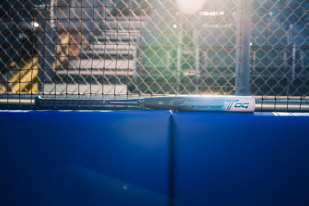 A silver and black 2025 Easton Ghost OG (-11) Fastpitch Softball Bat by Easton rests horizontally on a blue padded fence before a chain-link fence, with stadium seating in the background.