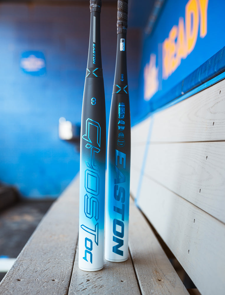 Two blue and white bats, including the 2025 Easton Ghost OG (-11) Fastpitch Softball Bat by Easton, lean against a wooden bench in a dugout with blue walls and blurred signage in the background.
