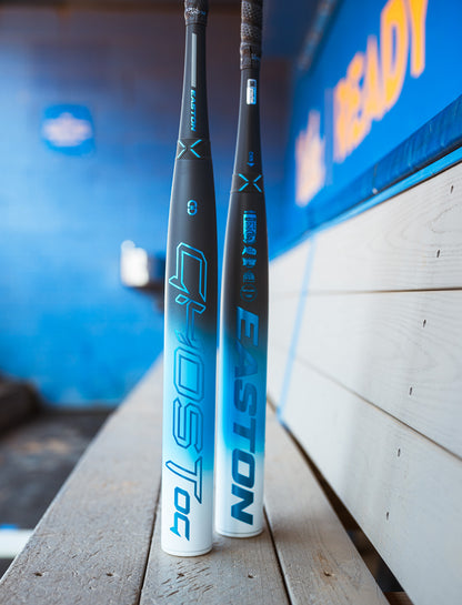 Two blue and white bats, including the 2025 Easton Ghost OG (-11) Fastpitch Softball Bat by Easton, lean against a wooden bench in a dugout with blue walls and blurred signage in the background.
