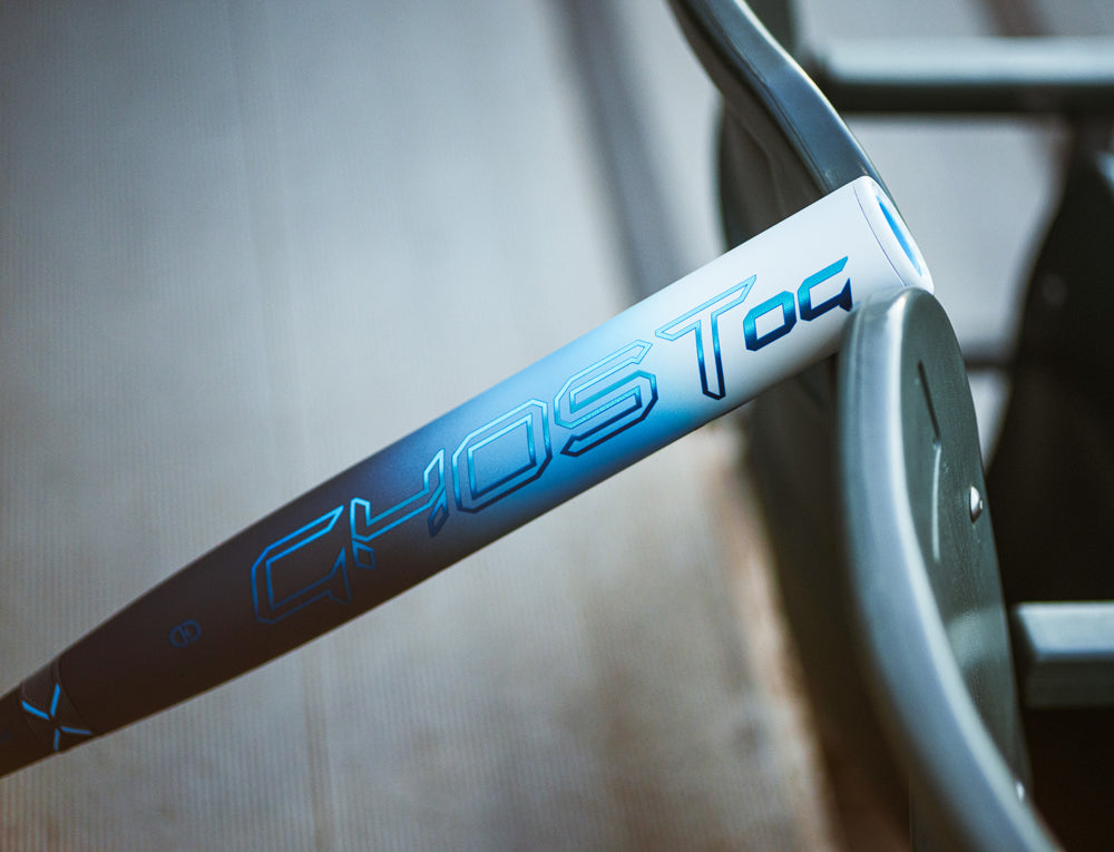 A close-up of the 2025 Easton Ghost OG (-11) Fastpitch Softball Bat (EFP5GHOG11) by Easton, showing the GHOST logo and Double Barrel design in blue and white, resting against a green metal fence.