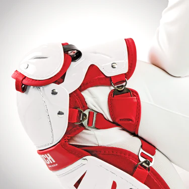 Close-up of a hockey goalie’s leg pad and knee guard in red and white, showing the detailed straps, buckles, and padding against a plain white background.