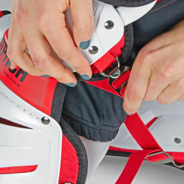 Close-up of a person with teal-painted nails fastening a red strap on white and red sports protective gear, likely hockey goalie pads, over black pants.