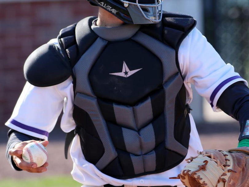 A baseball catcher wearing protective gear prepares to throw a baseball with their right hand while holding a glove in their left hand. The players face is partially obscured by a helmet and mask.