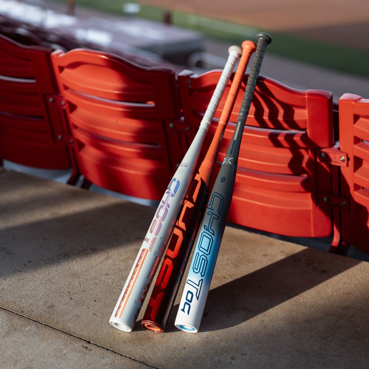 Two white 2026 Easton Ghost Advanced (-8) Fastpitch Softball Bats (EFP6GHAD8) with blue and orange GHOST logos lean against red stadium seats, their Double Barrel construction highlighted by the sunlight.