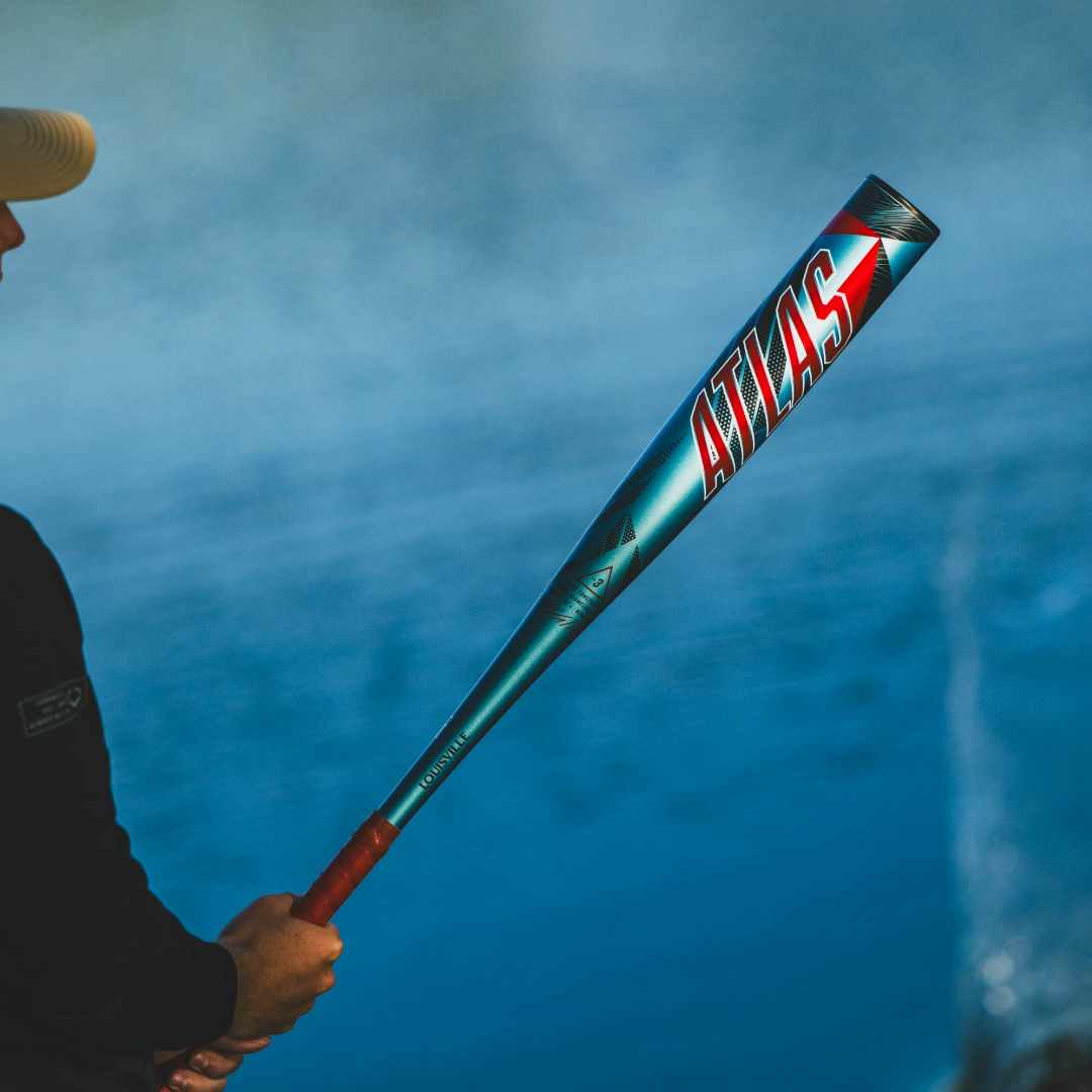 A person holds a metallic blue and red 2026 Louisville Slugger Atlas (-3) BBCOR Baseball Bat (WBL4116010) with "ATLAS" on the barrel, against a blurred background of water and mist.