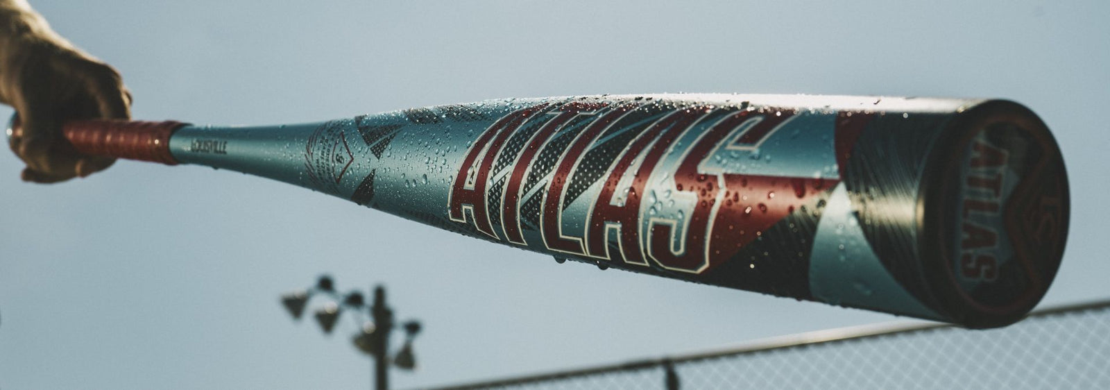 A close-up of a hand holding a wet metallic baseball bat labeled ATLAS, with water droplets visible and a blurred background of a fence, sky, and stadium lights.