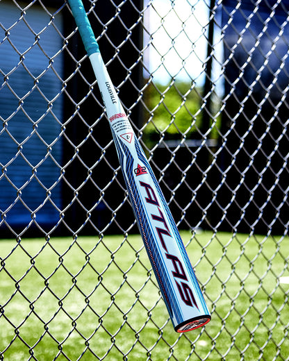 A white and blue Louisville Slugger 2027 Atlas 2.0 (-3) BBCOR Baseball Bat (WBL4248010), designed with EVX Alloy Technology for a balanced swing weight, leans against a chain-link fence with green grass and a blue building in the background.