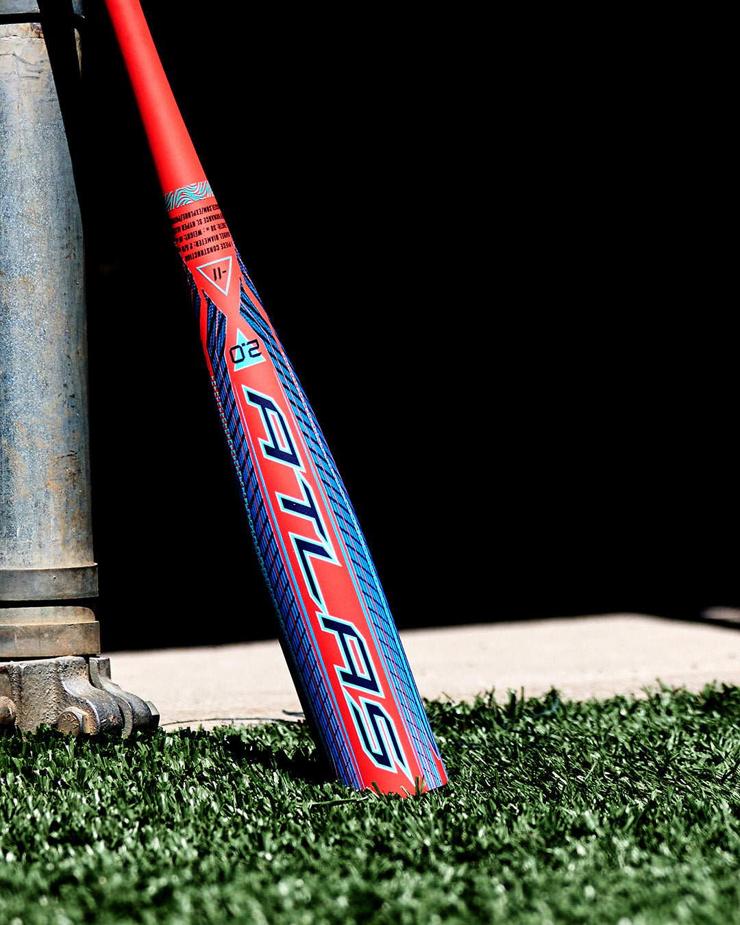 A 2027 Louisville Slugger Atlas 2.0 (-5) 2 5/8″ USA Baseball Bat (WBL4264010), featuring red and blue accents and a balanced swing weight, leans against a metal structure on artificial grass as sunlight casts nearby shadows.