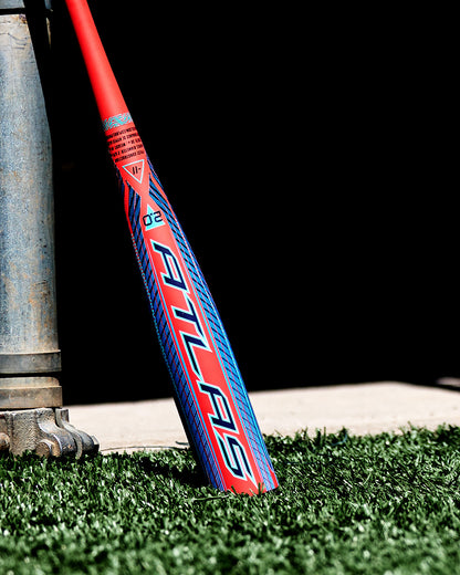 A 2027 Louisville Slugger Atlas 2.0 (-5) 2 5/8″ USA Baseball Bat (WBL4264010), featuring red and blue accents and a balanced swing weight, leans against a metal structure on artificial grass as sunlight casts nearby shadows.