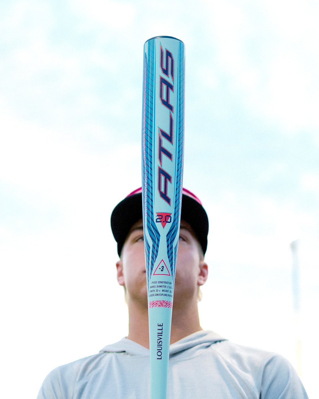 A person stands facing the camera, mostly hidden behind a blue and pink 2027 Louisville Slugger Atlas 2.0 (-3) BBCOR Baseball Bat (WBL4248010) held upright in front of their face against a bright sky background.