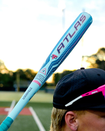 Wearing a black cap and pink sunglasses, a person stands on a baseball field at sunset holding the 2027 Louisville Slugger Atlas 2.0 (-3) BBCOR Baseball Bat (WBL4248010), known for its balanced swing weight, over their shoulder.