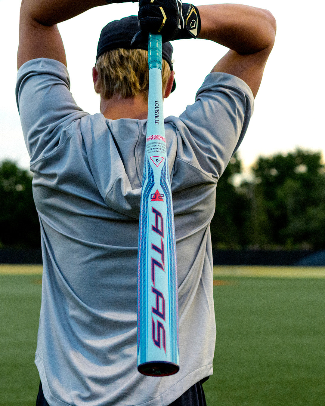A person wearing a gray shirt and black cap holds a blue and pink Louisville Slugger 2027 Atlas 2.0 (-3) BBCOR Baseball Bat (WBL4248010) over their shoulders on a baseball field with green grass and trees in the background, highlighting the bat’s balanced swing weight and advanced EVX Alloy Technology.