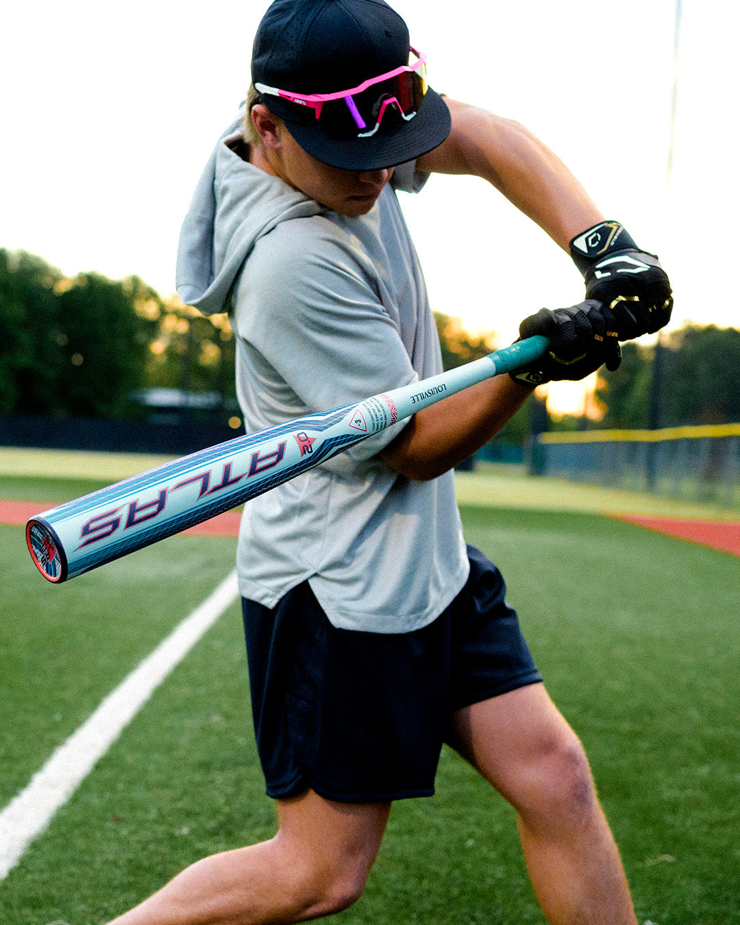 A baseball player wearing sunglasses and a hoodie swings the 2027 Louisville Slugger Atlas 2.0 (-3) BBCOR Baseball Bat (WBL4248010) on the field, showcasing its balanced swing weight with green grass and a fence in the background.