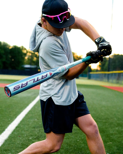 A baseball player wearing sunglasses and a hoodie swings the 2027 Louisville Slugger Atlas 2.0 (-3) BBCOR Baseball Bat (WBL4248010) on the field, showcasing its balanced swing weight with green grass and a fence in the background.