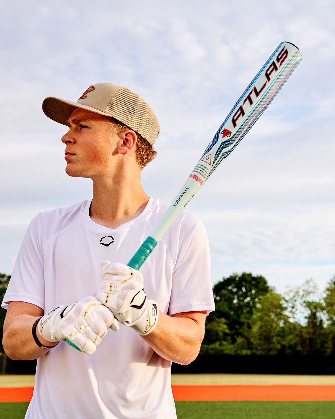 A baseball player wearing a white shirt, tan cap, and white gloves stands on a field with trees and sky in the background, holding a Louisville Slugger Atlas 2.0 (-3) BBCOR Baseball Bat (WBL4248010) on his shoulder.