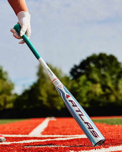 A person wearing a white glove holds the 2027 Louisville Slugger Atlas 2.0 (-3) BBCOR Baseball Bat (WBL4248010), which features EVX Alloy Technology and a balanced swing weight, while standing on a red baseball field with green trees and blue sky in the background.