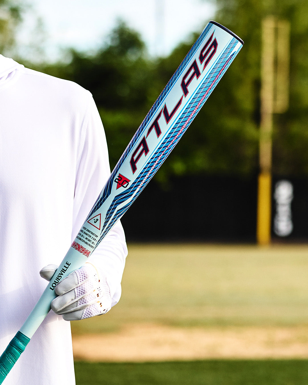 Wearing a white shirt and glove, a person holds the Louisville Slugger Atlas 2.0 (-3) BBCOR Baseball Bat (model WBL4248010) on a baseball field, showcasing its balanced swing weight with green trees and a yellow foul pole in the background.