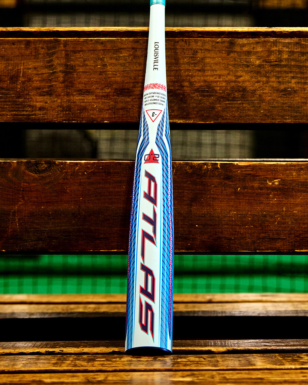 A 2027 Louisville Slugger Atlas 2.0 (-3) BBCOR Baseball Bat (model WBL4248010) with blue and red graphics stands upright on a wooden bench, featuring a balanced swing weight and advanced EVX Alloy Technology from Louisville Slugger, set against a blurred green and black background.