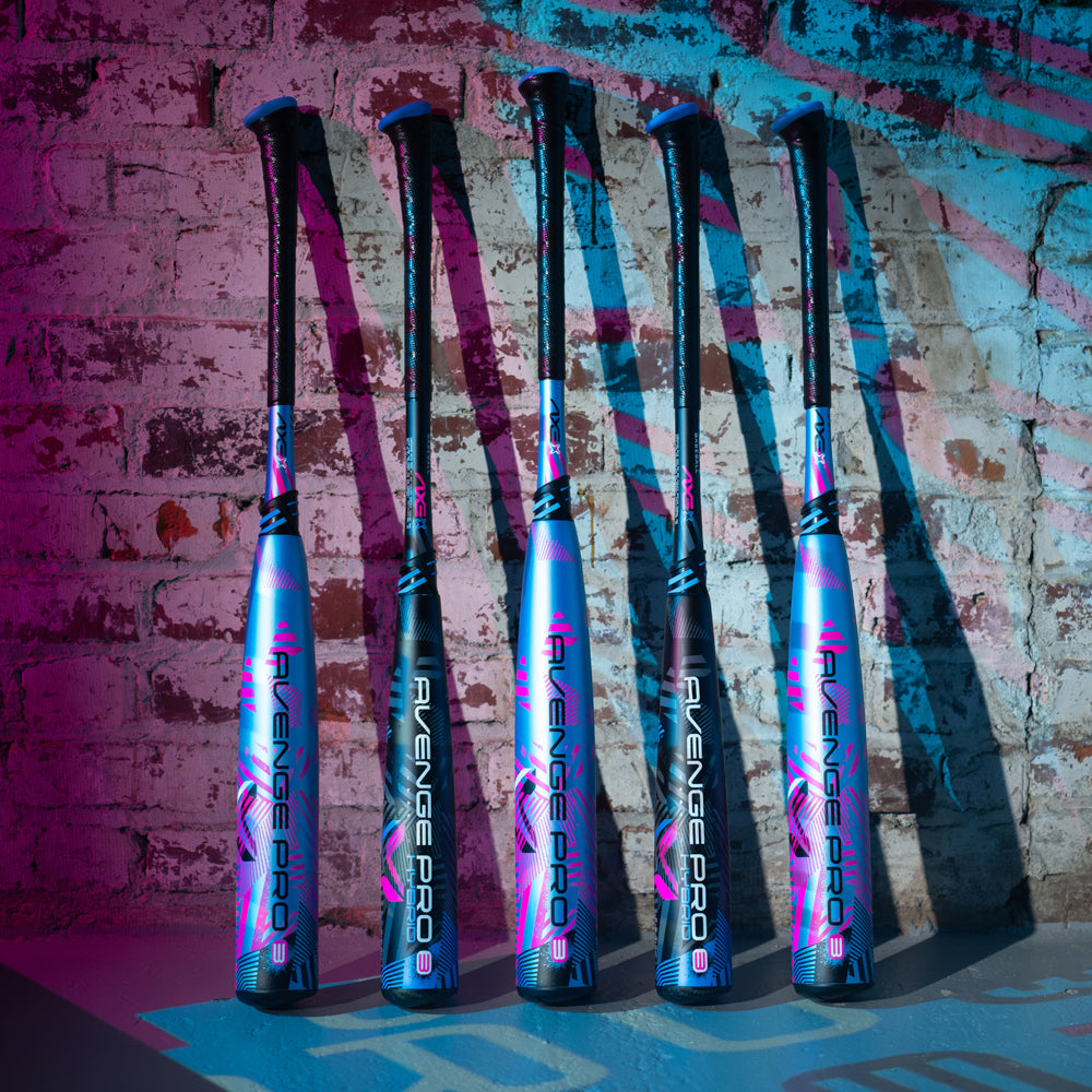 Five 2024 AXE Avenge Pro 3 Hybrid (-8) USA baseball bats with blue, pink, and black designs stand upright against a white brick wall, lit by blue and pink lights.