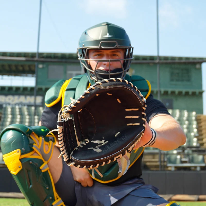 A baseball catcher in full gear squats on the field, holding an open All Star Pro-Elite Japan 34″ Baseball Catcher’s Mitt: CM3000-J1-34 made from premium HIMEJI leather toward the camera, with empty stadium seats and a green structure visible in the background.