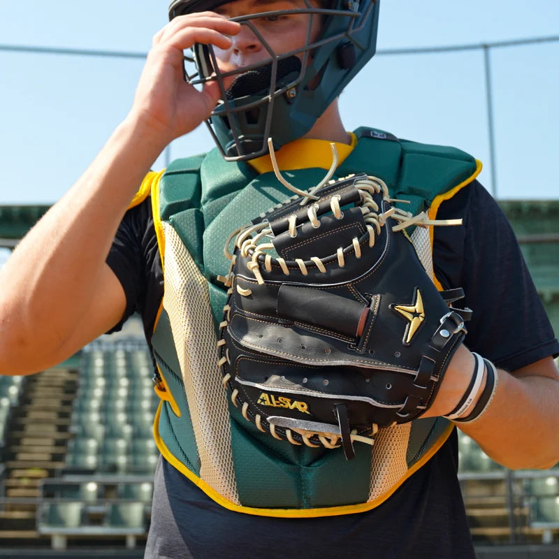 On a sunny day at an empty stadium, a catcher in protective gear adjusts his face mask while holding the All Star Pro-Elite Japan 34″ Baseball Catcher’s Mitt (CM3000-J1-34), a black mitt made from premium HIMEJI leather by All Star.