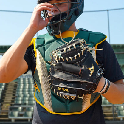 On a sunny day at an empty stadium, a catcher in protective gear adjusts his face mask while holding the All Star Pro-Elite Japan 34″ Baseball Catcher’s Mitt (CM3000-J1-34), a black mitt made from premium HIMEJI leather by All Star.