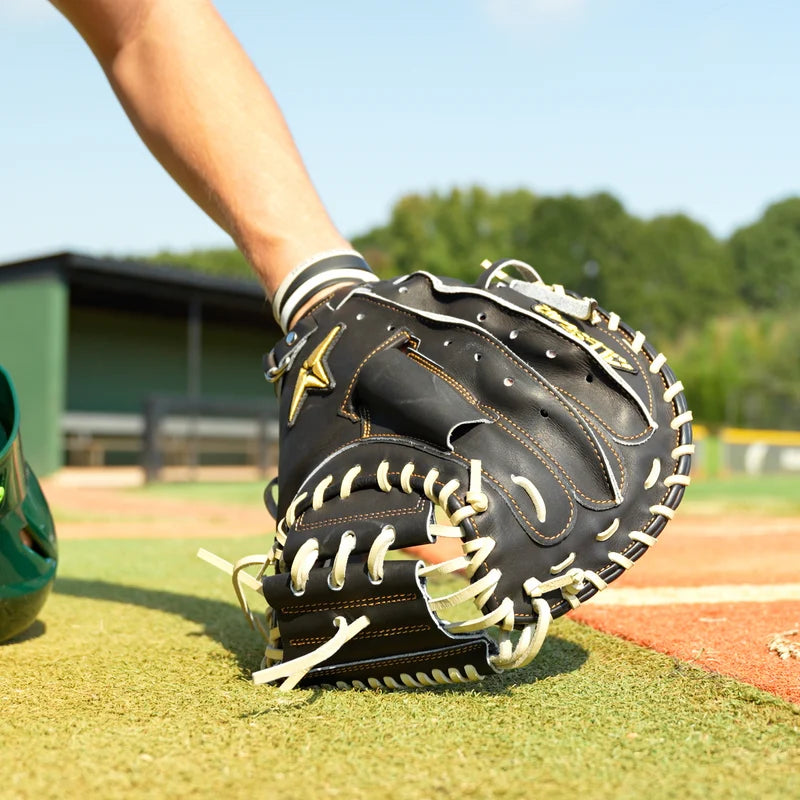 Wearing an All Star Pro-Elite Japan 34″ Baseball Catcher’s Mitt (CM3000-J1-34), a person crouches near home plate on a baseball field, with dugouts and trees in the background.