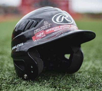 A black Rawlings baseball batting helmet rests on green artificial turf, displaying the brand logo and white text on the front and side. A sports field with blurred red and brown elements is visible in the background.