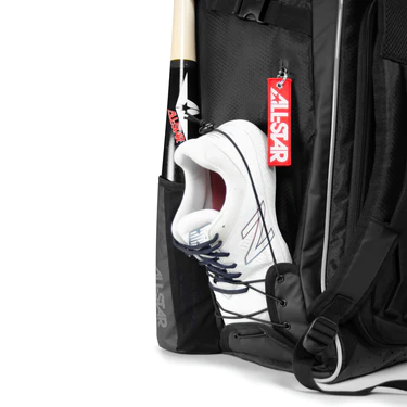 A close-up of an All Star MVP PRO Series Catcher's Backpack: BBCBP, black, with a pair of white athletic shoes and a baseball bat in the side pockets. A red "ALL-STAR" tag hangs from the bag.