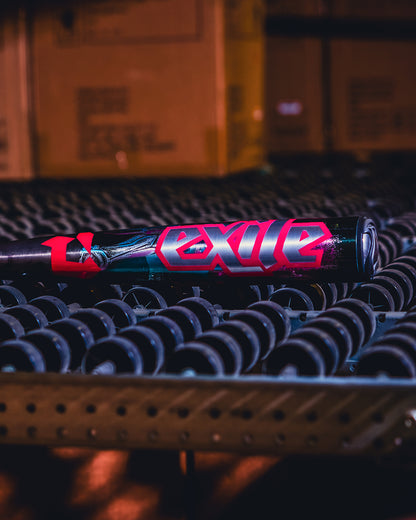 A 2026 DeMarini Exile Cyborg (-3) BBCOR Baseball Bat (WBD2654010), with striking EXILE graphics and X14 Alloy Barrel, sits on a roller conveyor in a warehouse as cardboard boxes blur in the background.