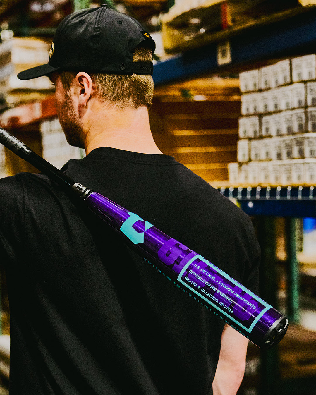 A man in a black shirt and hat holds a DeMarini 2026 The Goods Royal Purple (-3) BBCOR Baseball Bat (WBD2634010) over his shoulder, standing in a warehouse lined with shelves of boxes.