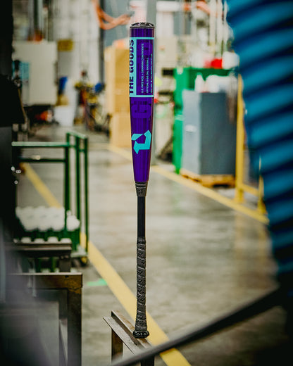 The 2026 DeMarini The Goods Royal Purple (-3) BBCOR Baseball Bat (WBD2634010) by DeMarini stands upright on a metal surface in an industrial warehouse, surrounded by blurred machinery and equipment.