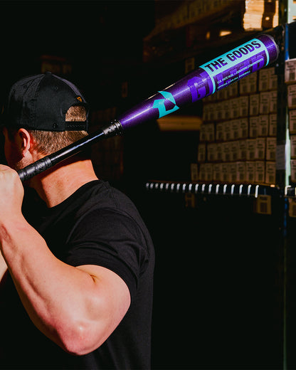 A person in a black shirt and cap holds the 2026 DeMarini The Goods Royal Purple (-3) BBCOR Baseball Bat: WBD2634010, featuring DeMarini branding, over their shoulder with stacked boxes in the background.