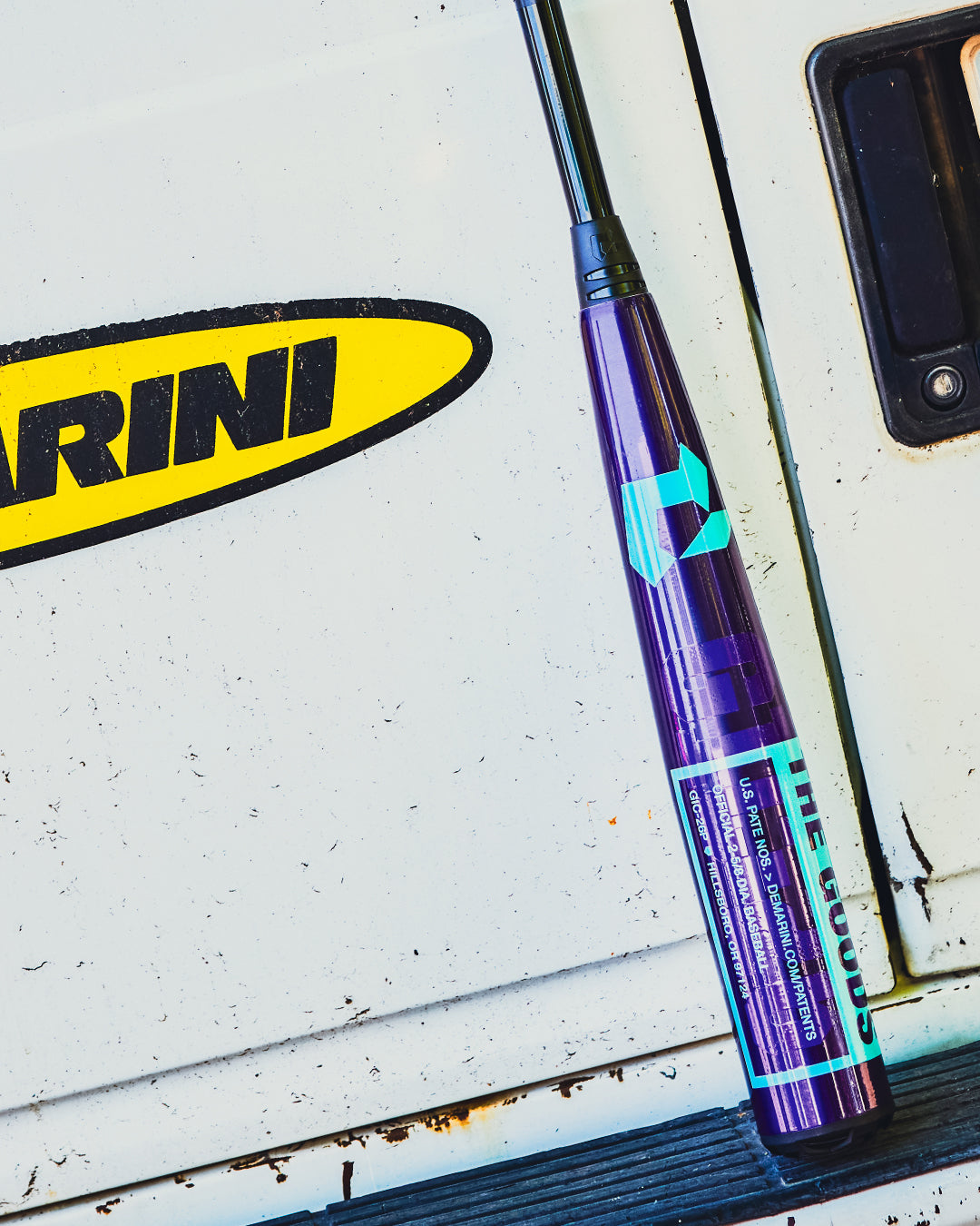 A 2026 DeMarini The Goods Royal Purple (-3) BBCOR Baseball Bat (WBD2634010) by DeMarini leans against a white vehicle door, with part of a yellow and black RINI sticker visible.